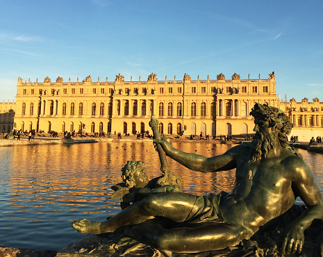 Paris, France - November, 2016: Gardens of Versailles. Statue on the Water Parterre at sunset. The palace facade can be seen in the background. This is one of the most visited monuments in France.