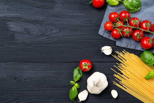 Food ingredients for spaghetti pasta with cherry tomatoes, fresh basil leaves and garlic, black wooden background, top view with copy space