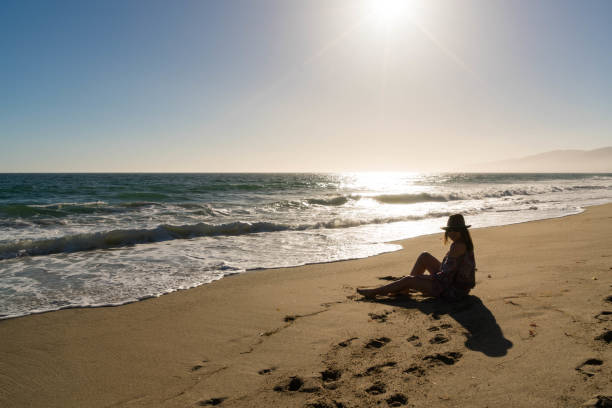 Woman Sits on Ocean Sand at Sunset A woman sits on sand at sunset. rock sea malibu silhouette stock pictures, royalty-free photos & images