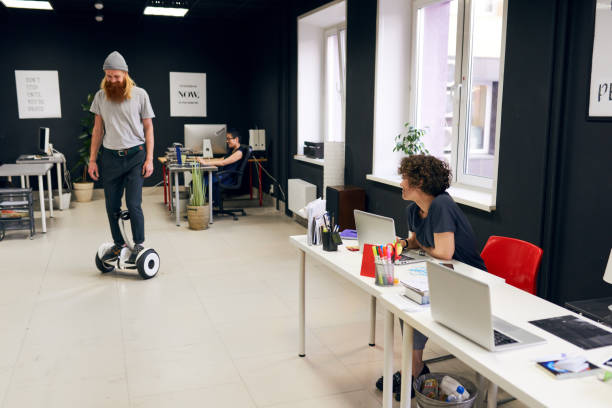 Modern office Jolly bearded hipster male manager in hat moving on hoverboard over open-plan office, curly-haired colleague looking at him and sitting at table hoverboard stock pictures, royalty-free photos & images