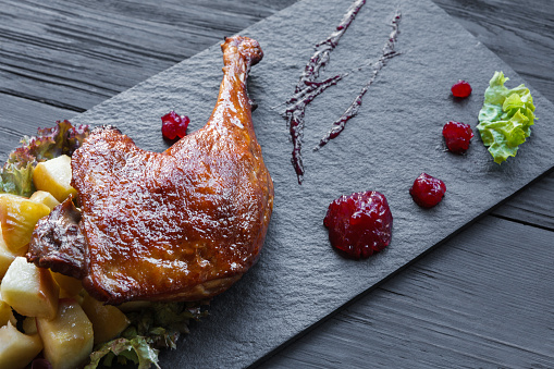 Roasted duck leg closeup, served on slate plate with apples, lettuce and cherry sauce. Restaurant food on black wood table