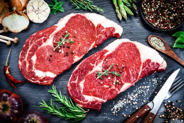 Top view of two fresh raw beef steak fillets ready for cooking shot on dark kitchen table surrounded by many cooking and seasoning ingredients. Predominant colors are red and black. Low key DSRL studio photo taken with Canon EOS 5D Mk II and Canon EF 100mm f/2.8L Macro IS USM