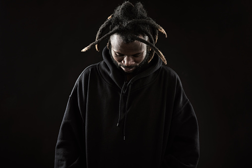 Melancholy young black man in sweatshirt with hood looking down and holding hands in pockets
