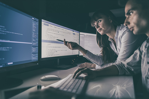 A woman and a man sit at a desk in a dimly lit room, focused on multiple computer screens displaying lines of code and programming information, with the woman gesturing towards the screen on the left.