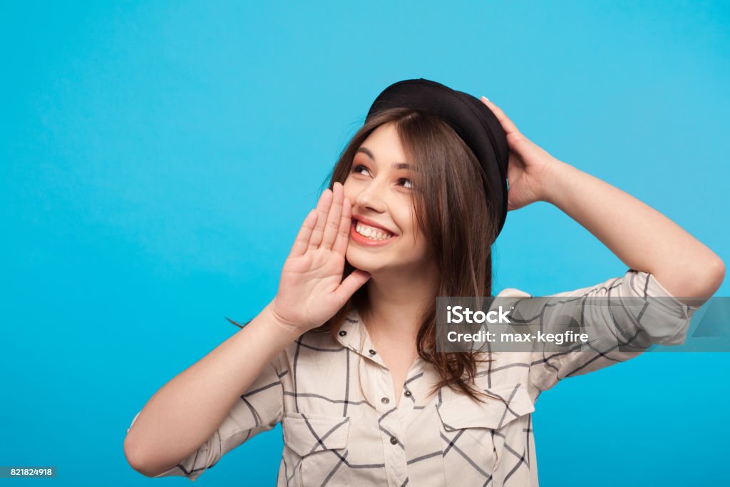 Trendy girl gossiping on blue background Young girl in shirt and hat holding hand on face telling secrets on blue background. Adult Stock Photo