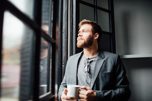 Indoor shot of thoughtful young businessman standing by window with a cup of coffee and looking outside.