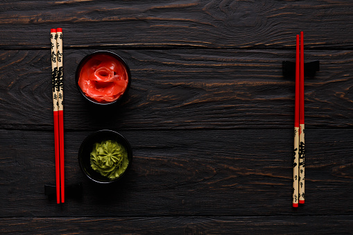 Pieces of traditional japanese tablewear. Red chopsticks with Bon appetite written in hierogliphs, small bowls with ginger and wasabi on dark wooden table. Top view, flat lay, copy space, background