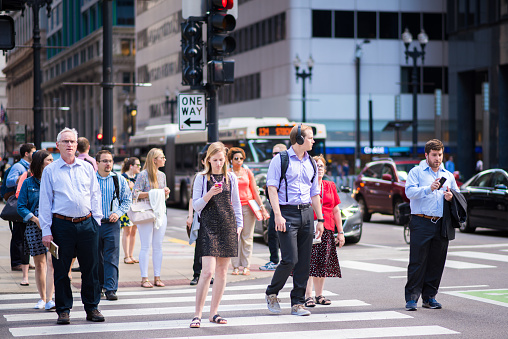 Chicago, IL, June 15, 2017: Stylish young woman checks her phone while waiting to cross the street, downtown. Cell phones are ubiquitous in the city, and people sometimes text while they walk.