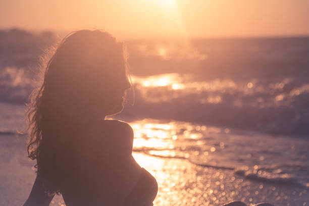 Silhouette portrait of young female sitting on beach against ocean waves and sunset over sea. Summer vacation concept.