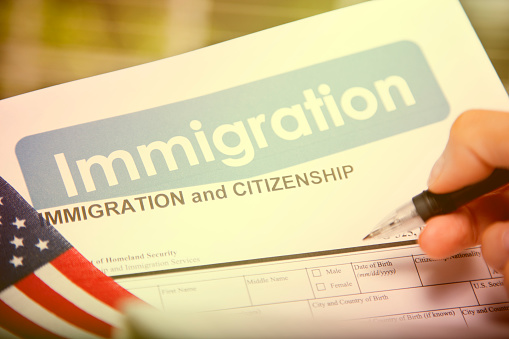 Person filling out an application for USA immigration, citizenship forms, paperwork with American flags on desk.  Close-up of hand, pen.