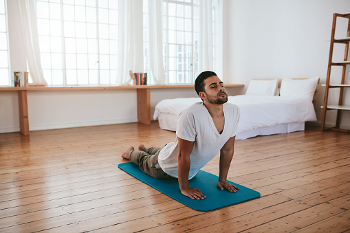 Indoor shot of handsome young man practicing yoga. Fitness man meditating with his eyes closed while doing cobra pose in living room.