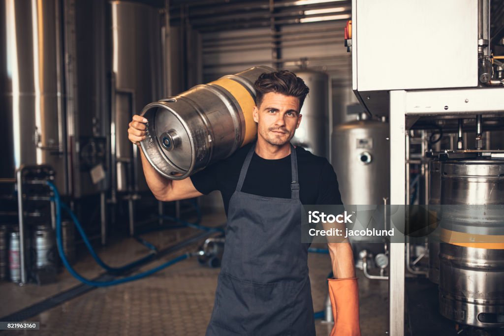 Male brewer carrying metal container at brewery factory Portrait of male brewer carrying metal container at brewery factory. Young man holding a keg on shoulder at warehouse. Brewery Stock Photo