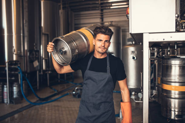 Male brewer carrying metal container at brewery factory Portrait of male brewer carrying metal container at brewery factory. Young man holding a keg on shoulder at warehouse. keg stock pictures, royalty-free photos & images