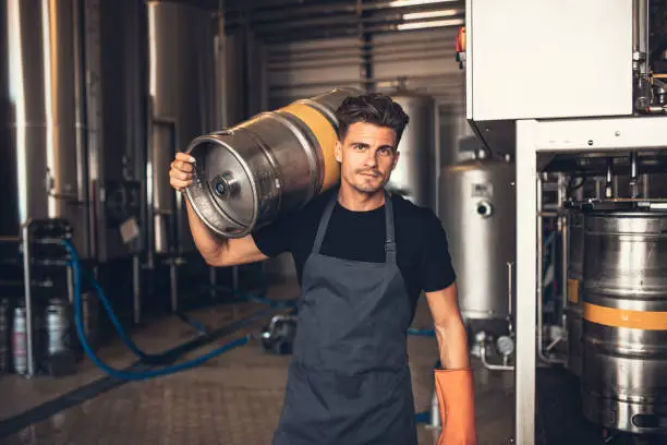 Portrait of male brewer carrying metal container at brewery factory. Young man holding a keg on shoulder at warehouse.