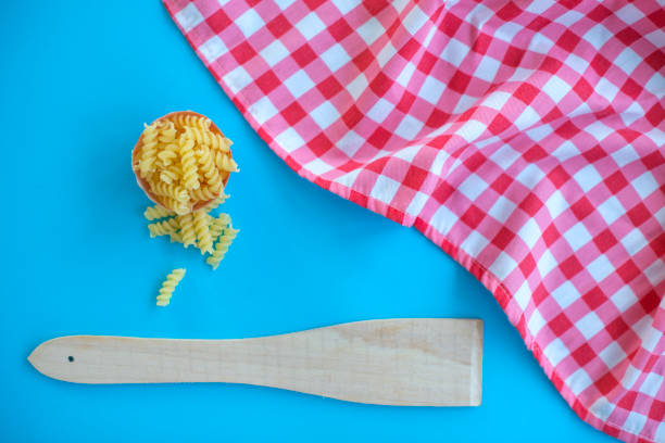 Pasta, wooden spatula and checkered tablecloth on blue background.