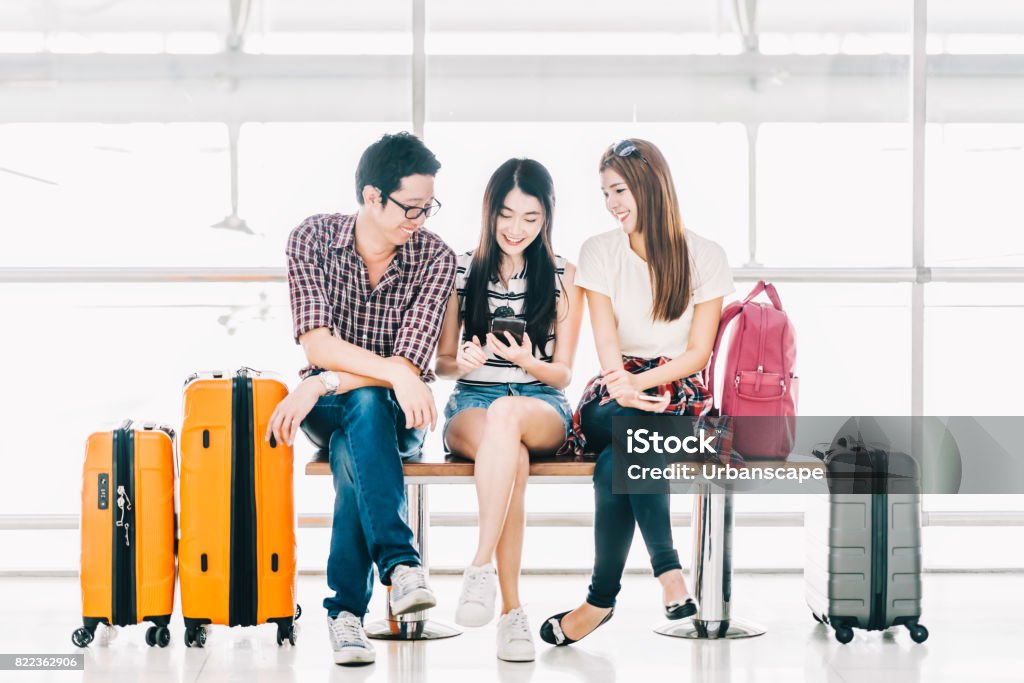 Group of young Asian travelers using smartphone checking flight or online check-in at airport together, with luggage. Travel abroad, summer holiday trip, or mobile phone application technology concept Tourist Stock Photo