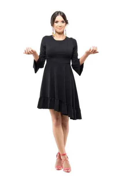 Smiling charming uncertain woman in black dress shrugging shoulders with raised arms. Full body length portrait isolated on white background.