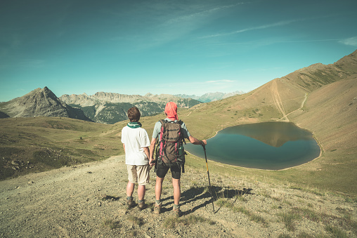 Couple of hiker on the mountain top looking at blue lake and mountain peaks. Summer adventures on the Alps. Wide angle view from above, toned image, vintage style.