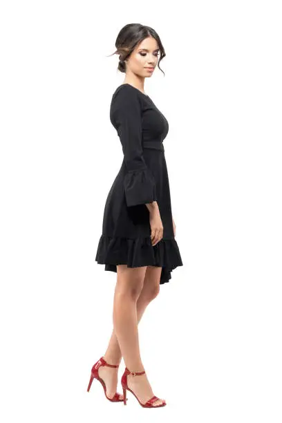 Side view of young woman in black dress standing and looking down. Full body length portrait isolated on white background.