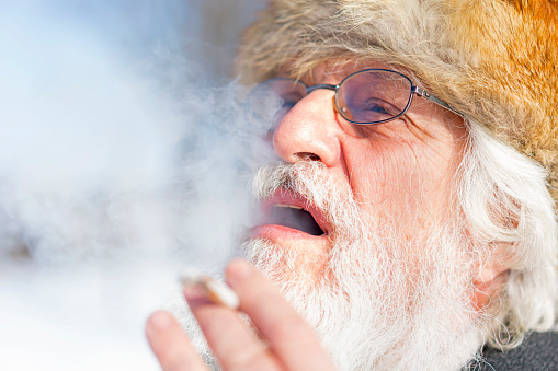 Portrait of senior with white beard, wears a bitch or a fur hat, glasses and is exhaling smoke while smoking a cigarette.