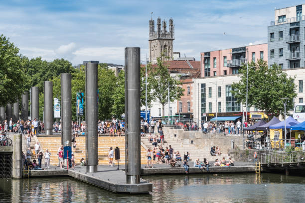 The Waterfront area of Bristol with boats moored up. People walking along the quay and sitting in the sun.
