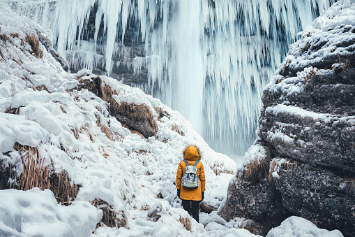 Woman with orange jacket standing below amazing frozen Pericnik waterfall (Slovenia).