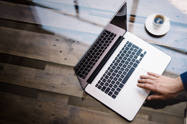 istock Man using laptop at sunny office.Male hand touching notebook trackpad.Modern notebook, cup of black coffee on the wooden table.Reflections on glass surface.Vertical.Top view. 822690554