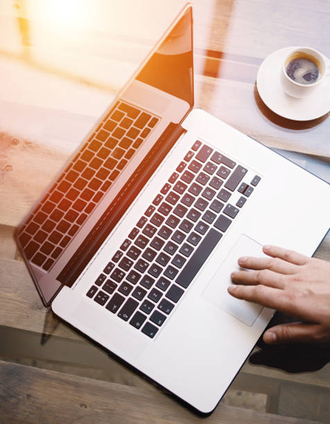 Man working with laptop at sunny office.Male hand pointing on notebook trackpad.Modern notebook, cup of black coffee on the wooden table.Reflections on glass surface.Vertical.Top view.Visual effect.