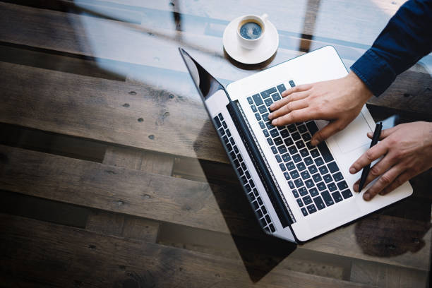 istock Man working on laptop at sunny office.Male hand typing on keyboard.Modern notebook, cup of black coffee on the wooden table.Reflections on glass surface.Vertical.Top view. 822691812