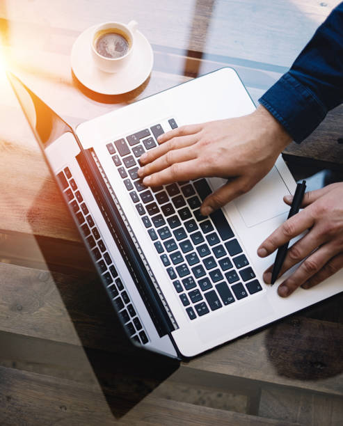 istock Man working on laptop at sunny office.Male hand typing on keyboard.Modern notebook, cup of black coffee on the wooden table.Reflections on glass surface.Vertical.Top view.Visual effect. 822691944
