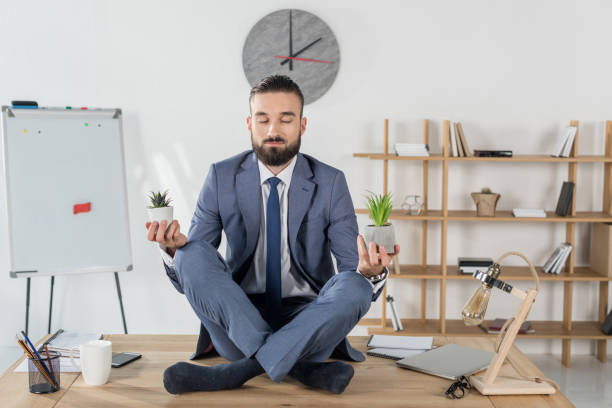 istock businessman with eyes closed meditating in lotus position while sitting on table in office 822692508