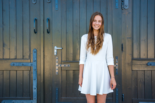 Young teenage girl standing in white dress in front of dark wooden door ready to enter
