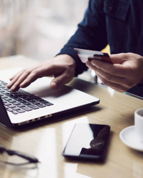 homem de negócios, mantendo o cartão de crédito de plástico na mão e usando o computador portátil enquanto está sentado na mesa de madeira. homem fazendo compras on-line. fundo desfocado. vertical. - credit card e commerce security finance - fotografias e filmes do acervo