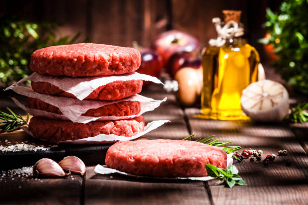 Raw burger patties on rustic wooden table Front view view of raw patties for making burgers shot on rustic wooden kitchen table. Some cooking and seasoning ingredients like rosemary, garlic, peppercorns, olive oil and oregano are on the table. Predominant colors are red and brown. Low key DSRL studio photo taken with Canon EOS 5D Mk II and Canon EF 70-200mm f/2.8L IS II USM Telephoto Zoom Lens spanish hamburger stock pictures, royalty-free photos & images