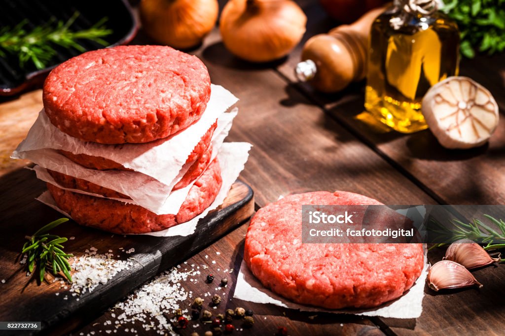 Raw burger patties on rustic wooden table High angle view of raw patties for making burgers shot on rustic wooden kitchen table. Some cooking and seasoning ingredients like rosemary, garlic, peppercorns, olive oil and oregano are on the table. Predominant colors are red and brown. Low key DSRL studio photo taken with Canon EOS 5D Mk II and Canon EF 70-200mm f/2.8L IS II USM Telephoto Zoom Lens Raw Food Stock Photo