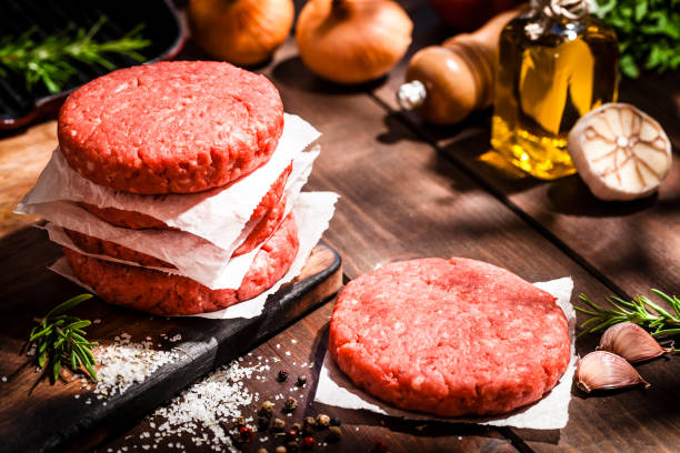 Raw burger patties on rustic wooden table High angle view of raw patties for making burgers shot on rustic wooden kitchen table. Some cooking and seasoning ingredients like rosemary, garlic, peppercorns, olive oil and oregano are on the table. Predominant colors are red and brown. Low key DSRL studio photo taken with Canon EOS 5D Mk II and Canon EF 70-200mm f/2.8L IS II USM Telephoto Zoom Lens ground beef photos stock pictures, royalty-free photos & images