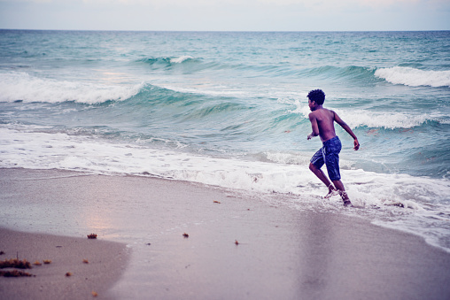 Young african american boy lost in his world playing by himself with the ocean waves at dusk. Purple and blue tone. Horizontal full length outdoors shot with copy space. This was taken in Florida, USA.