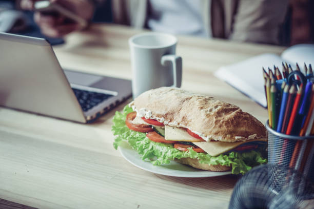 Close up view of working palce of freelancer during lunch. Wooden desk with laptop, cup of coffee and sandwich on it.
