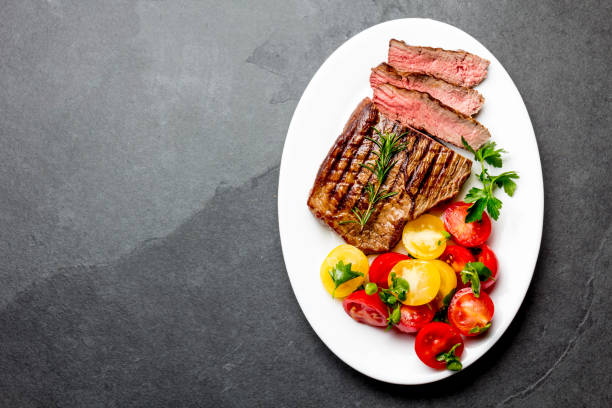 istock Sliced medium rare grilled beef steak served on white plate with tomato salad and potatoes balls. Barbecue, bbq meat beef tenderloin. Top view, slate background, copy space 823187284