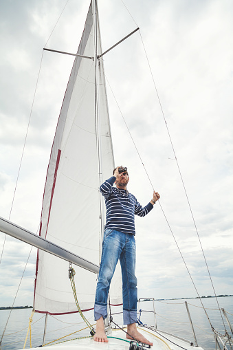 Barefoot man in a striped shirt and jeans looking through binoculars sailing yacht on a background cloudy sky