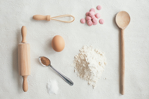 Kitchen ingredients and utensils on white background: rolling pin, wire whisk, four, egg, spoons and chocolate.
