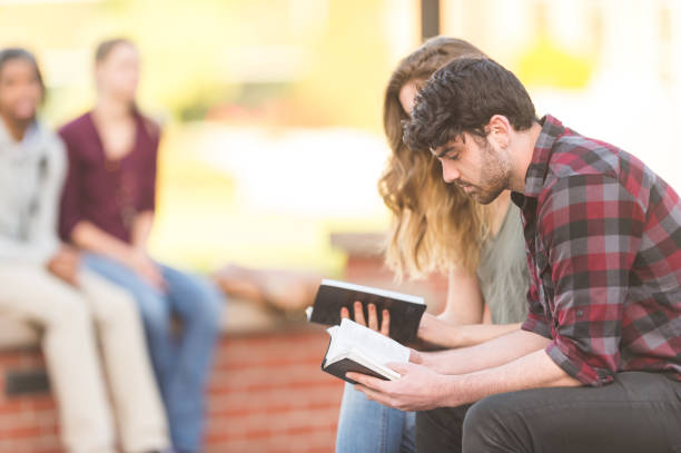 Bible Study Two college students - a male and female - study and read  the Bible together on a park bench outside. religious text stock pictures, royalty-free photos & images