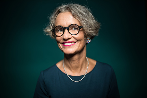 Portrait of cheerful woman in eyeglasses against green background. Smiling female is wearing jewelry. She is having short gray hair.