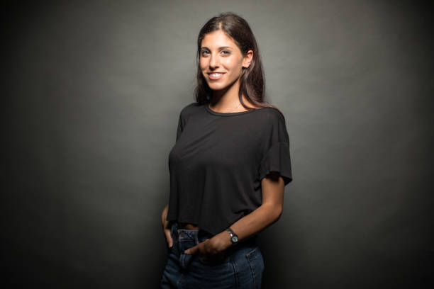 Happy Young Woman Standing With Hands In Pockets Portrait of cheerful young woman with hands in pockets. Beautiful female is wearing casuals. She is standing against gray background. three quarter length stock pictures, royalty-free photos & images