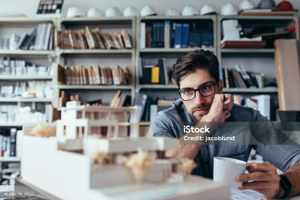 Thoughtful male designer looking at model with cup of coffee Thoughtful male designer in office looking at model with cup of coffee. Young male architect taking break from work. Architectural Model Stock Photo