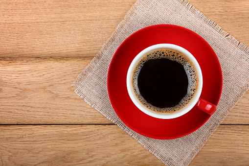 Full big cup of black instant coffee with froth on red porcelain saucer over wooden table with textile tablecloth napkin, close up, elevated top view, directly above