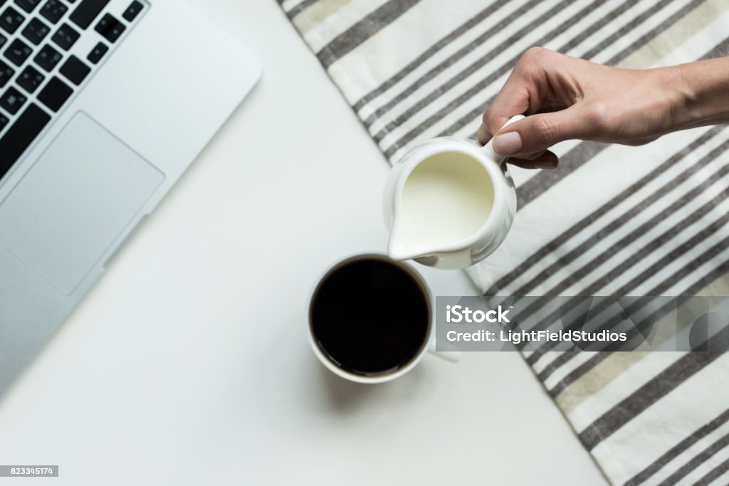 Person pouring milk from jug into cup of black coffee on white working desk Black Coffee Stock Photo