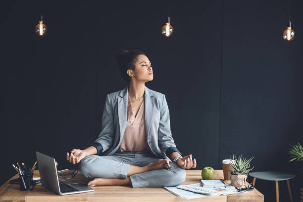 empresaria afroamericana sentada en postura de loto en mesa mientras meditaba en la oficina - con las piernas cruzadas fotografías e imágenes de stock