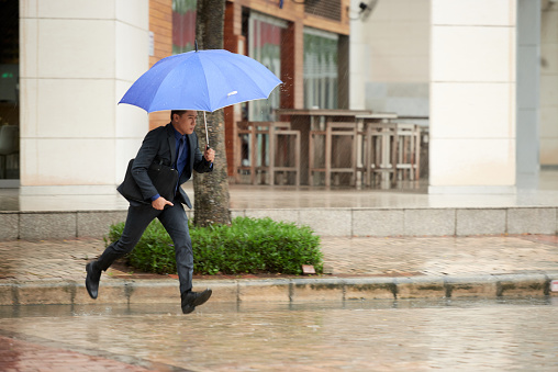 Portrait of businessman running fast across street in rain, holding umbrella