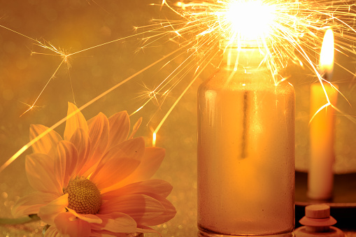 Concept of sparklers in Glass jar at night and fresh yellow flower with blurred golden light candle burning background.